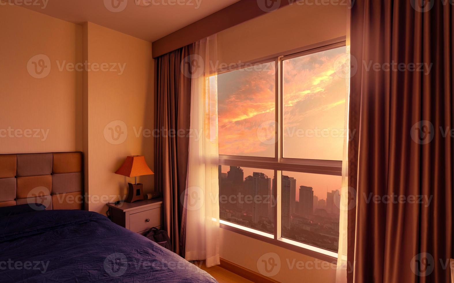 dormitorio por la mañana con la luz del sol por la mañana a través de una ventana de cristal con cortinas abiertas. cama de lujo en apartamento moderno en la ciudad. interior del dormitorio. cama cubierta con una manta azul. vista desde la ventana de cristal. foto