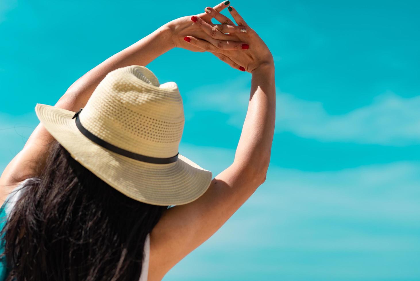 Back view of happy young Asian woman in casual style fashion and straw hat relax on holiday .Woman with green and red color nail manicure stretch arms against blue sky on summer vacation. Summer vibe. photo