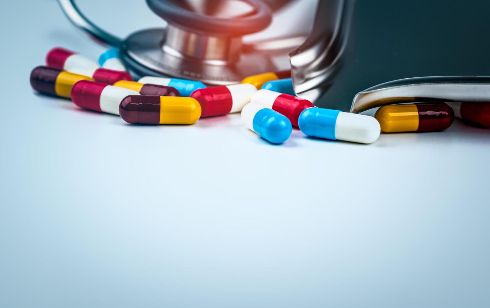 Stethoscope with pile of colorful antibiotic capsule pills on white table with drug tray. Antimicrobial drug resistance and overuse. Medical equipment for doctor. Global healthcare. Pharmaceuticals. photo