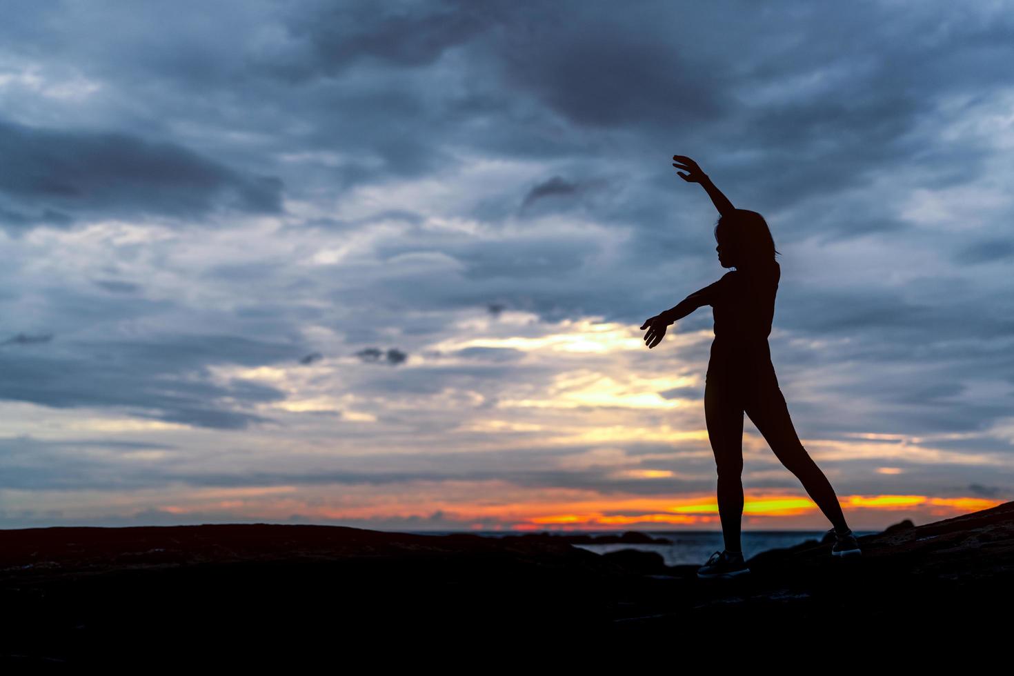 Silhouette woman workout in the morning at stone beach with beautiful sunrise sky. Fit woman stretching body before workout. Exercise for healthy lifestyle. Out door workout. Nature landscape. Freedom photo