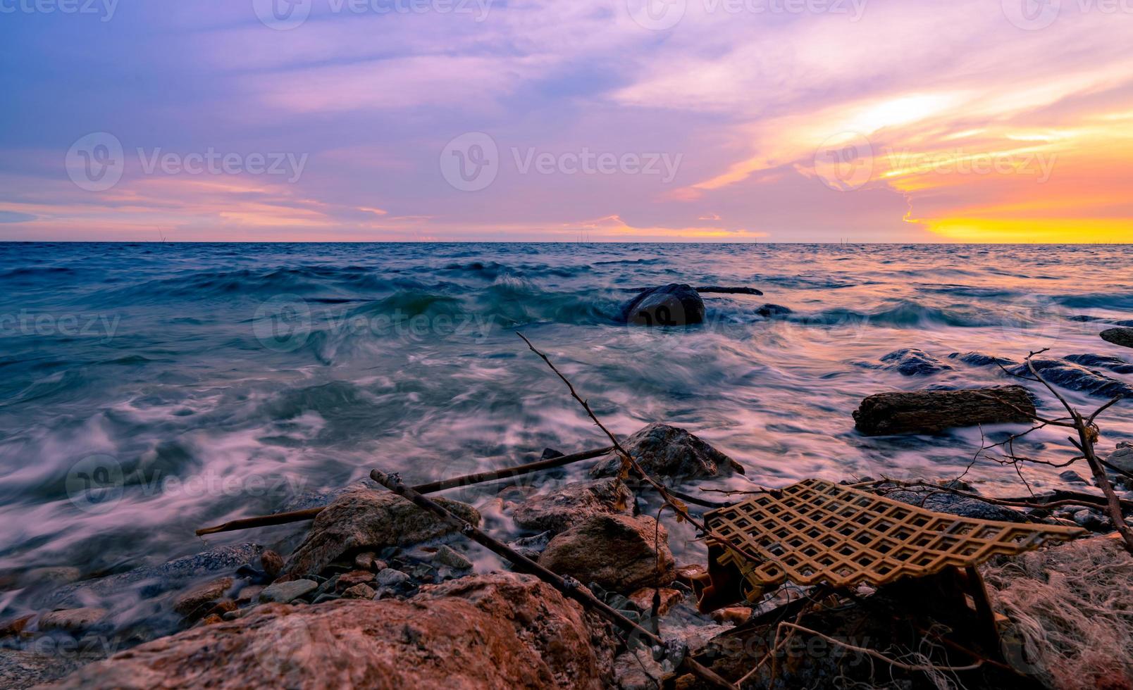 Sea water with golden and purple sunset sky. Plastic waste on rock beach. Tropical beach and ocean pollution. Ocean water. Seashore on summer. Soft wave at dusk. Seascape. Beauty in nature. photo