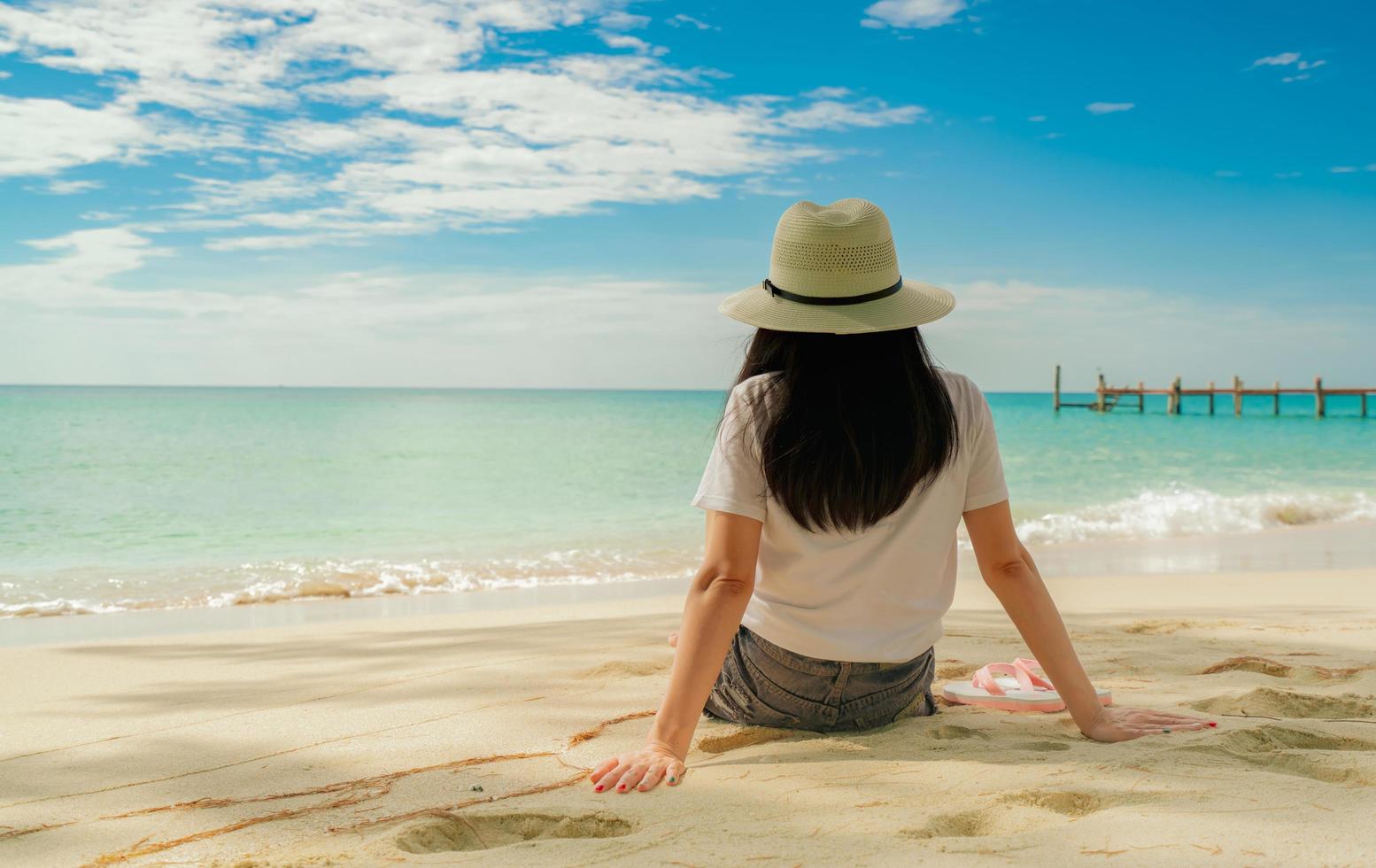 mujer joven feliz en camisas blancas y pantalones cortos sentados en la playa de arena. relajarse y disfrutar de unas vacaciones en la playa del paraíso tropical con cielo azul y nubes. chica en vacaciones de verano. vibras de verano. día feliz. foto