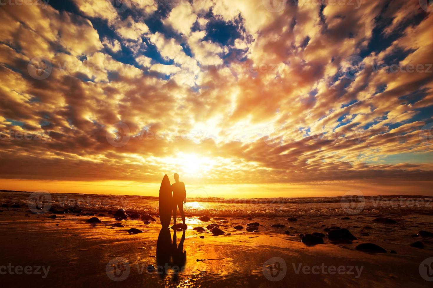 Surfer standing with his surfboard on the beach at sunset over the ocean photo