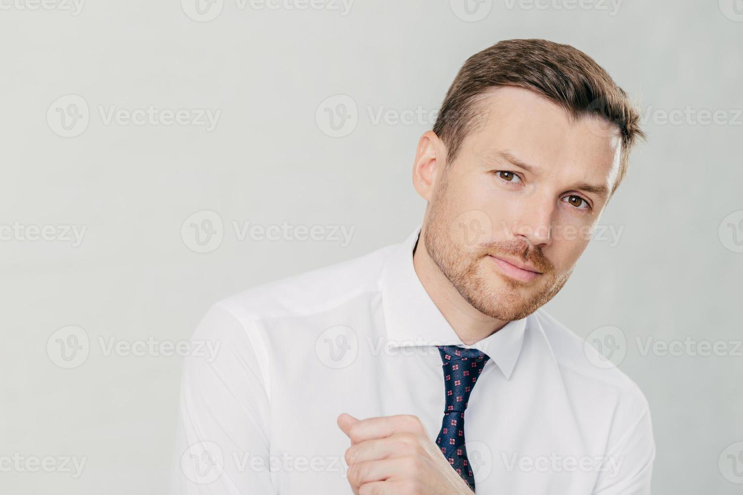Horizontal shot of handsome young male entrepreneur in formal white shirt, looks directly at camera, listens attentively his interlocutor, talk on business theme, isolated over white background photo