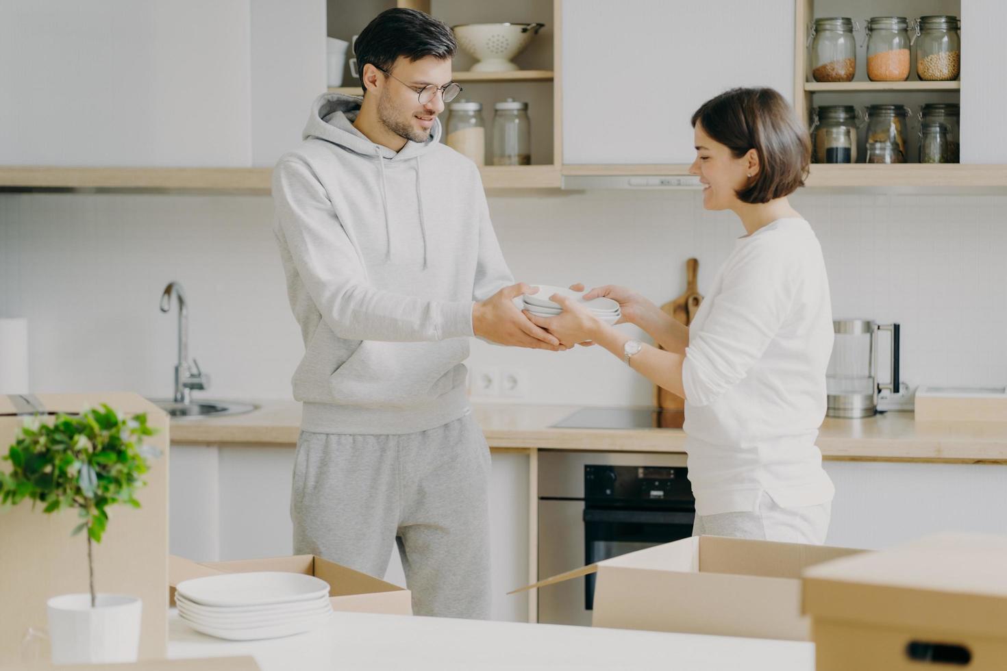 Horizontal shot of smiling couple unpack cardboard boxes in new kitchen, man passes plates to woman, placing dishes in cupboard, surrounded with moving packages in own bought or rented apartment photo