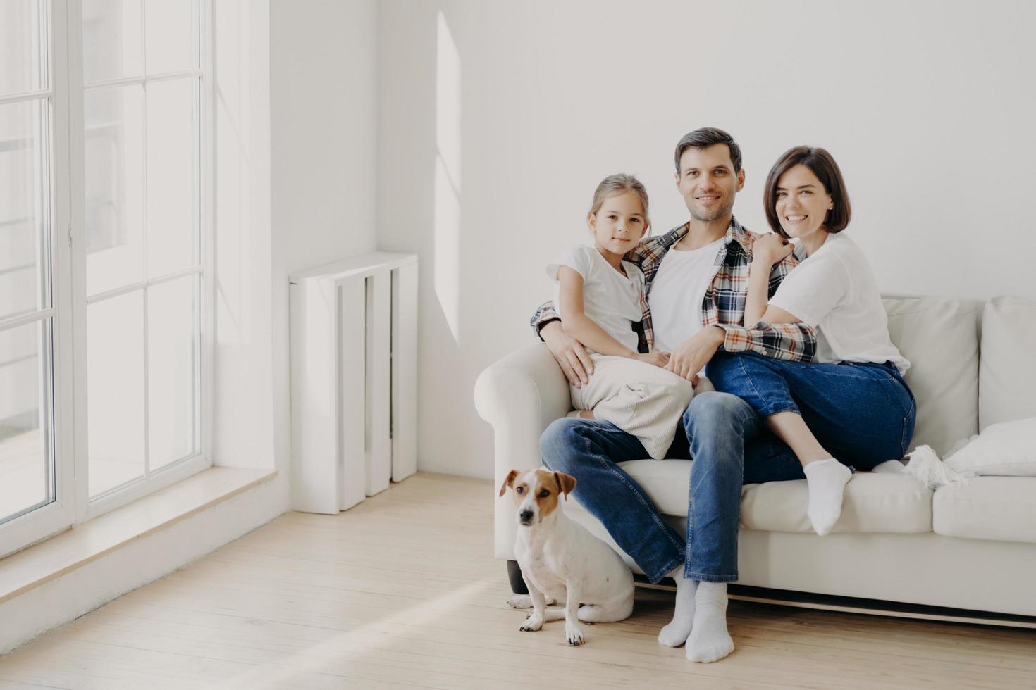 Family, togetherness and relationnship concept. Happy man embraces daughter and wife, sit on comfortable white sofa in empty room, their pet sits on floor, make family portrait for long memory photo