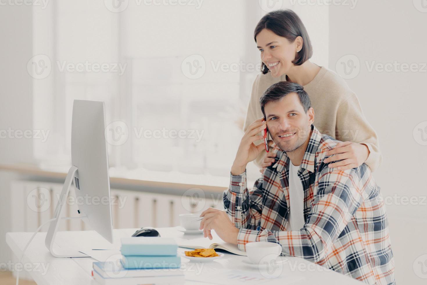 Happy husband and wife do paperwork together, pay taxes online look at screen computer, pose at desktop with books and papers, drink coffee and have lunch, make telephone call via smartphone photo