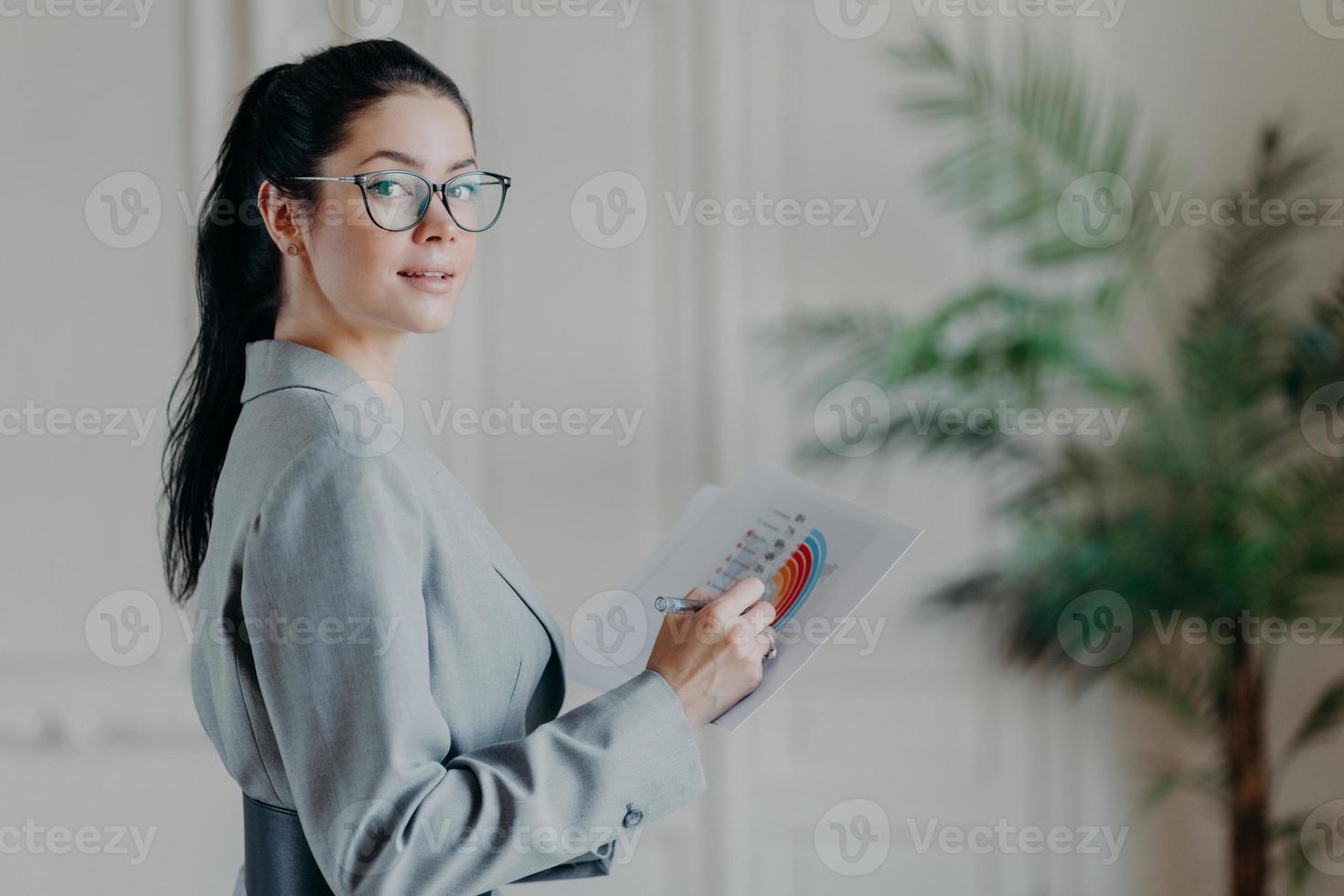 Horizontal shot of female strartuper analyzes business information, holds paper documents with diagrams and graphics, looks confidently at camera, wears transparent glasses and formal clothes photo