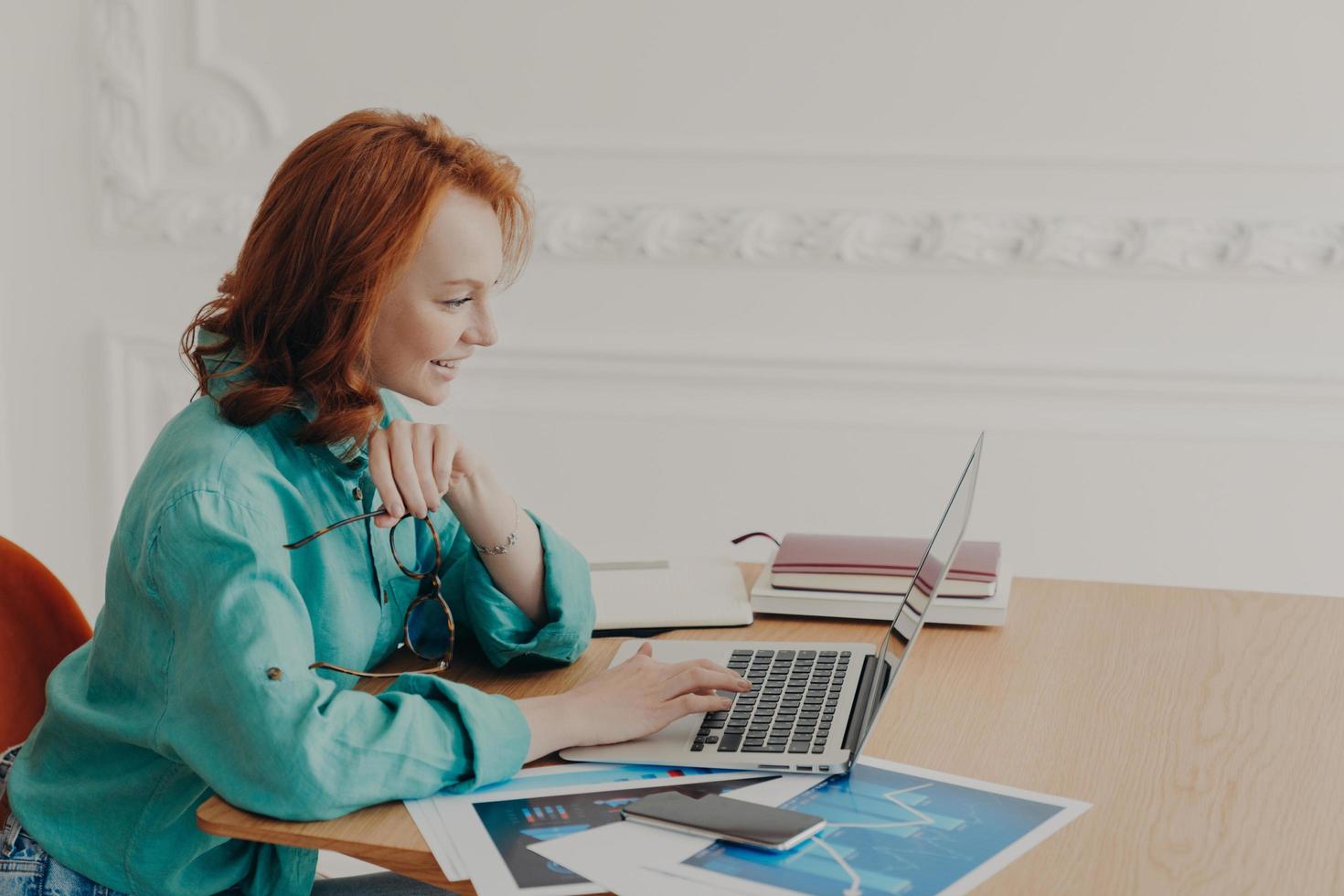 Female business owner prepares startup project, browses internet on laptop, works freelance, poses in workplace at home, does distant job, chats with colleagues has pleased expression dressed casually photo