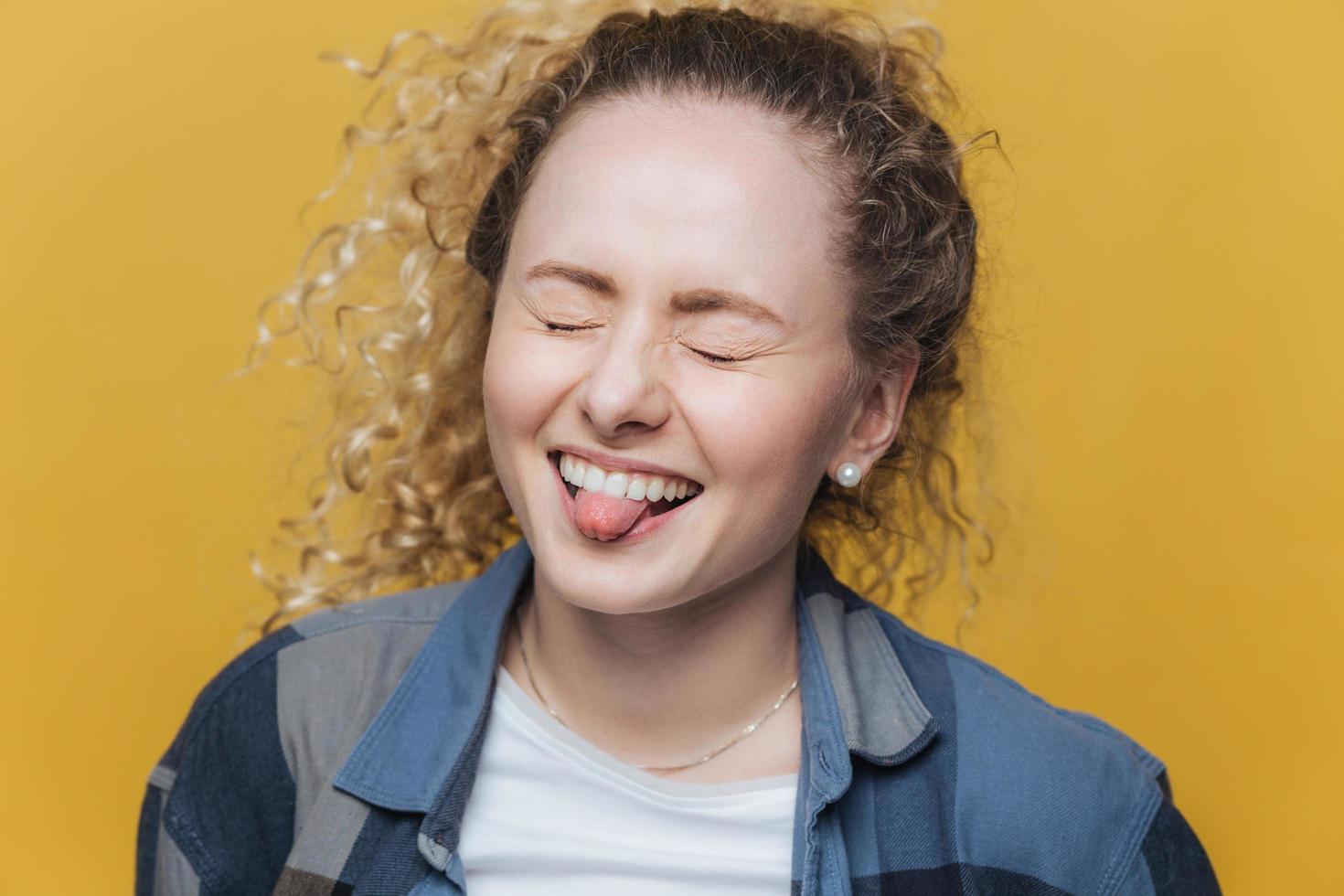 Photo of funny overjoyed female has fun alone, laughs joyfully and shows tongue, keeps eyes shut, expresses postiveness, isolated over yellow background. People and facial expressions concept