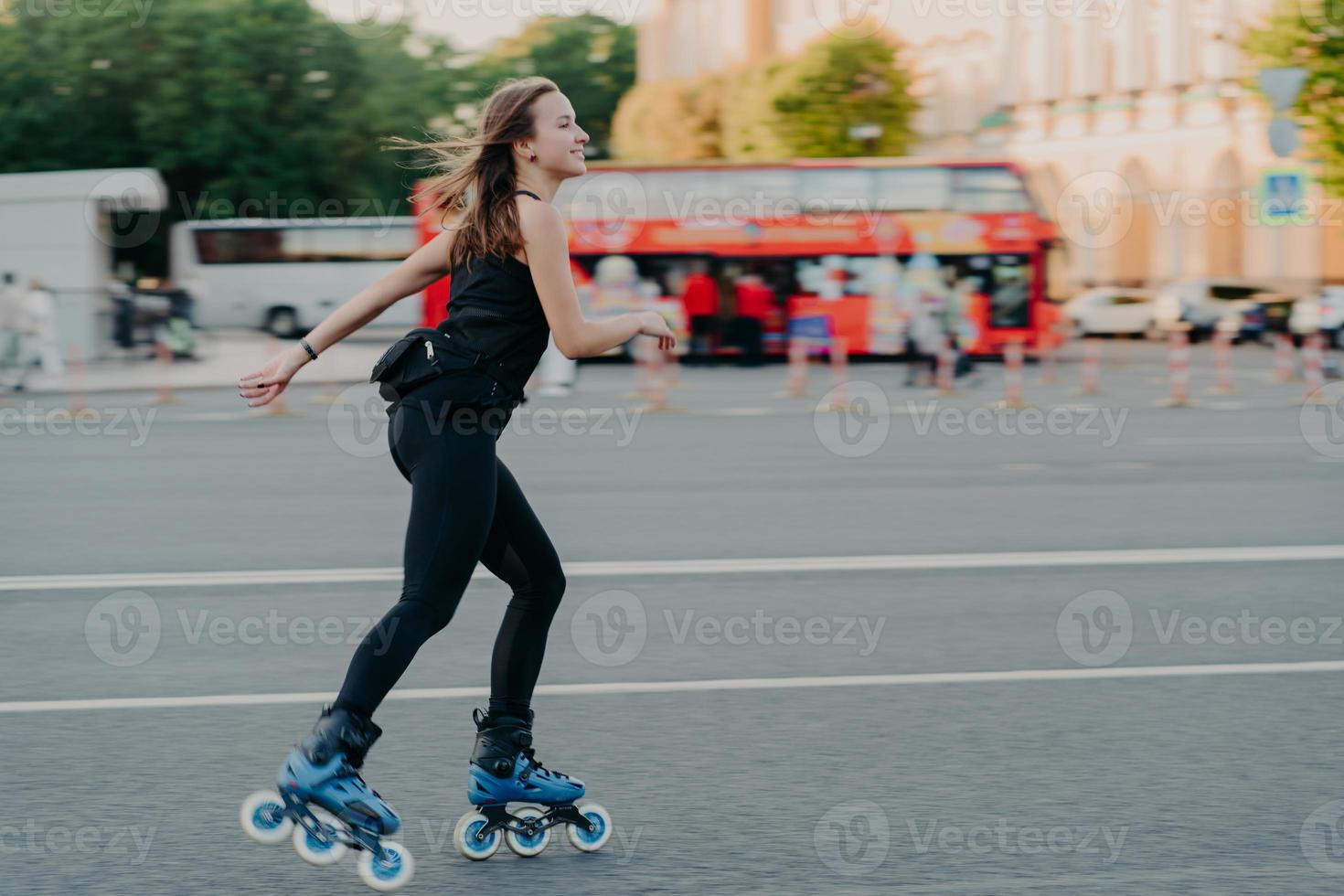 Outdoor shot of active slim young woman enjoys rollerskating during spare time dressed in blackactive wear poses in urban place on road against blurred background with transport. Hobby concept photo
