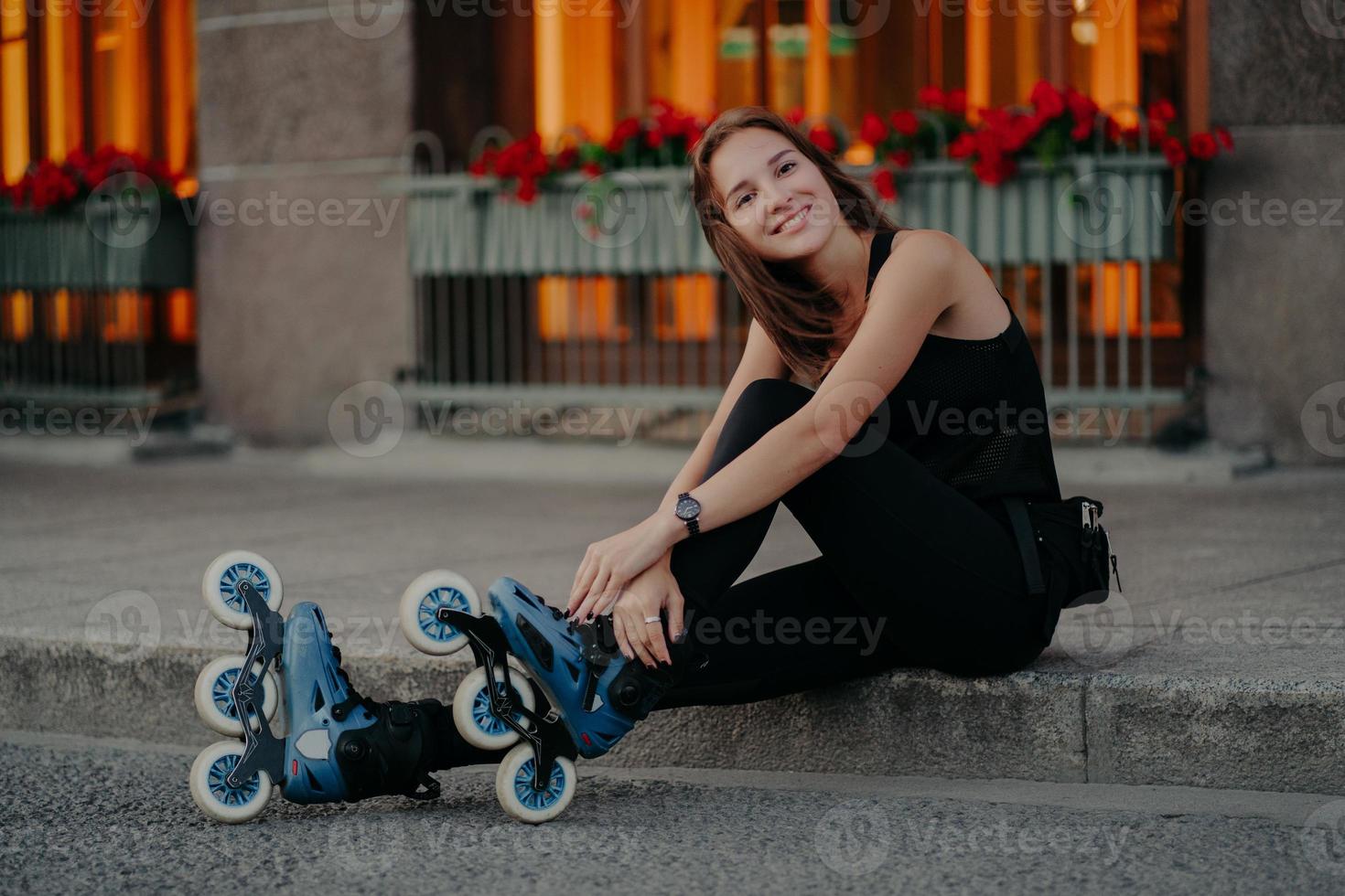 Una joven europea de aspecto agradable que usa patines se toma un descanso después de montar poses al aire libre vestida con ropa deportiva negra y tiene una sonrisa feliz en la cara. concepto de hobby y estilo de vida de recreación de personas foto