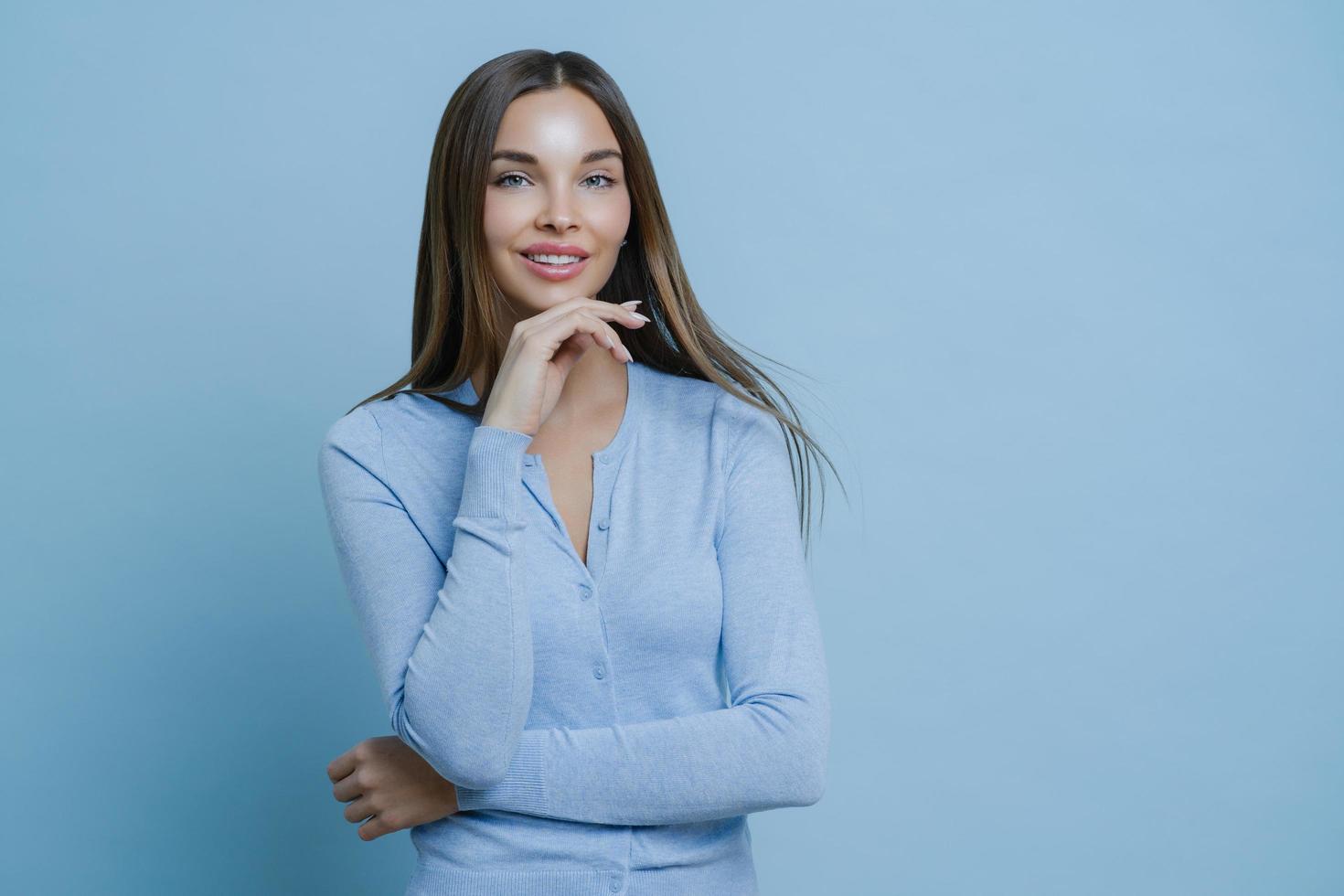 Feminity, beauty, wellness concept. Satisfied brunette lady touches jawline gently, has applied makeup, looks gently at camera with smile, thinks up great idea for personal business, wears blue jumper photo