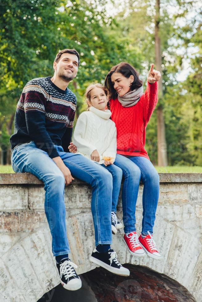 Carefree restful woman, man and small cute girl sits on bridge outdoors, admire sunrise. Affectionate mother shows to her little daughter beauty of nature. Family enjoys spending free time in park photo