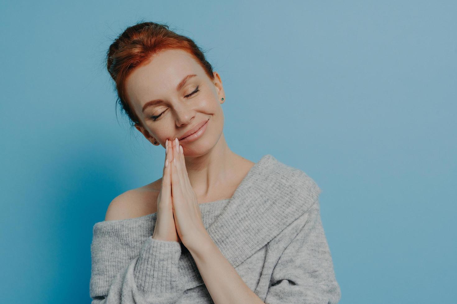 Happy calm redhead grateful woman holding hands in prayer gesture with closed eyes in studio photo