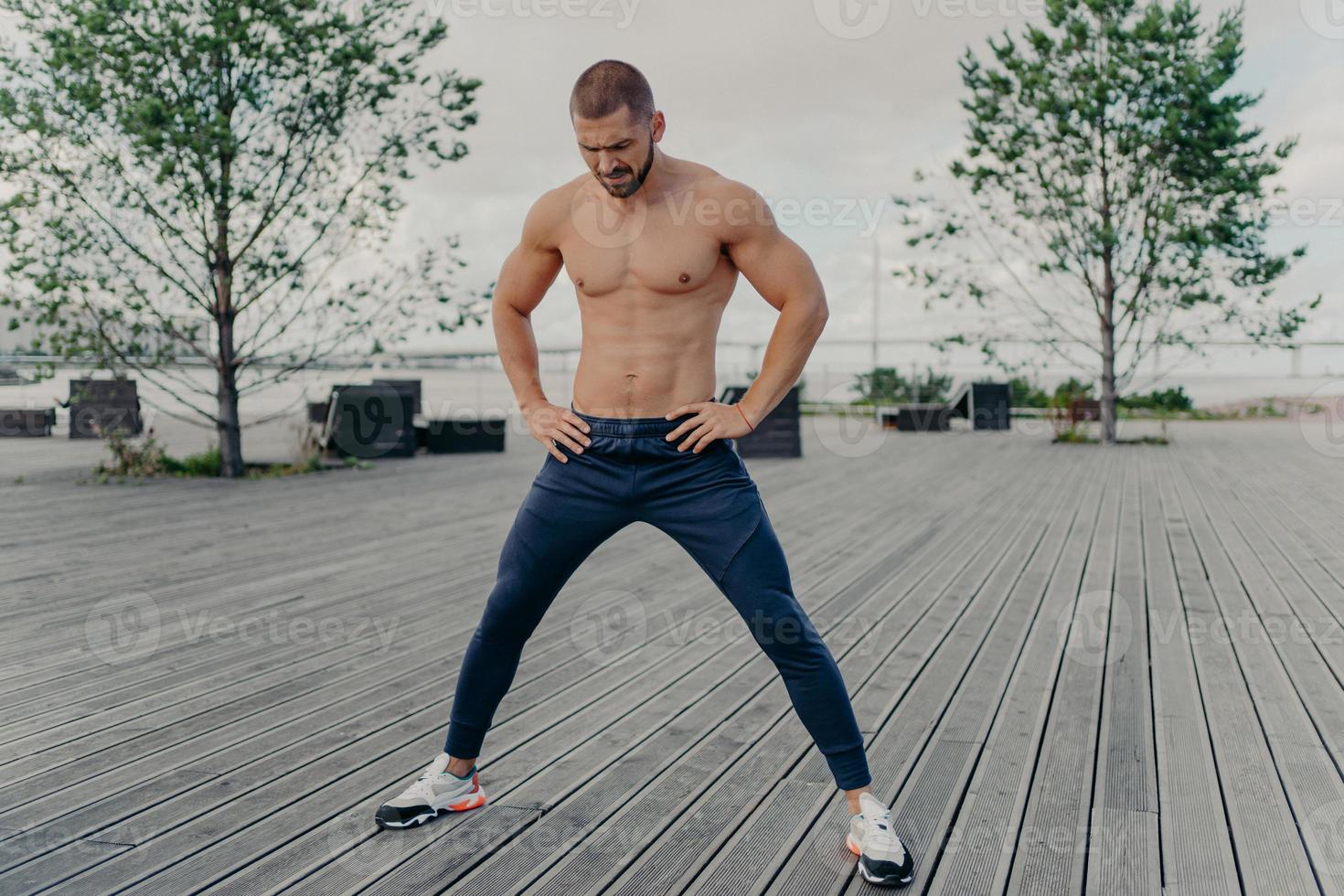 Outside of athlete guy does sport exercises in fresh air, stands with naked torso, has muscular body and stands outside. Active unshaven guy in good physical shape, has regular workout in morning photo