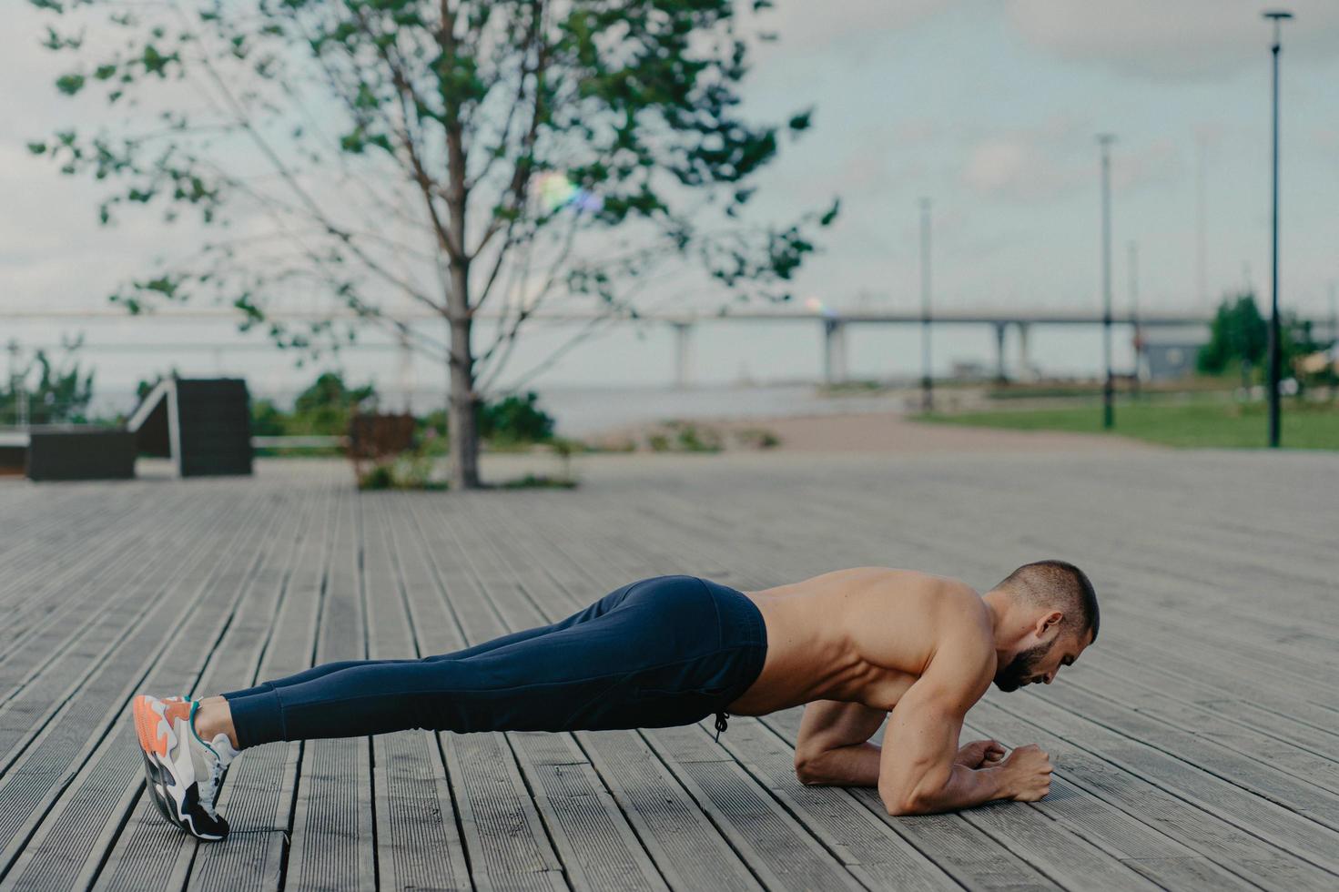 Horizontal shot of sporty bearded man stands in plank pose, practices yoga outdoor and dressed in active wear, breathes fresh air. Sport, fitness and healthy lifestyle concept. Training workout photo