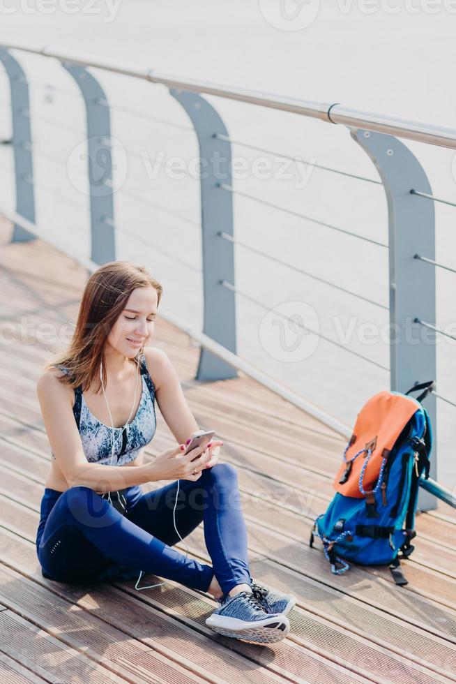 toma vertical de una atractiva joven activa que sostiene un teléfono celular moderno, navega por las redes sociales, escucha música con auriculares, disfruta del tiempo libre durante el fin de semana, recibe un mensaje de su mejor amiga foto