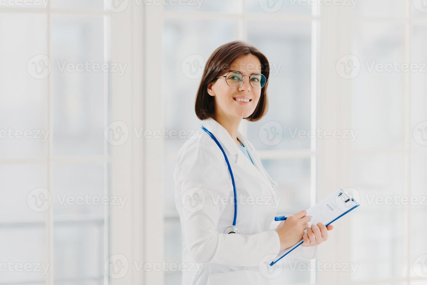 Horizontal shot of female doctor fills up medical form at clipboard, stands indoor, wears round glasses, white gown and stethoscope. General practitioner writes down notes, consults patients photo