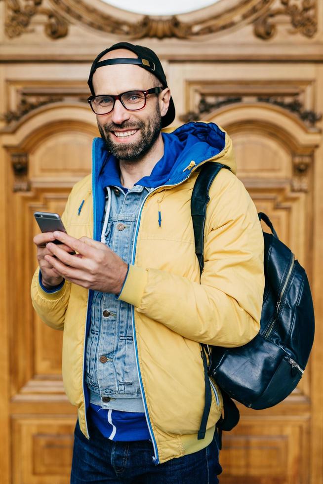 Hipster tourist in glasses, cap and yellow anorak holding backpack and smartphone having excursion in art gallery making photos being happy and delightful. People, tourism, technology concept