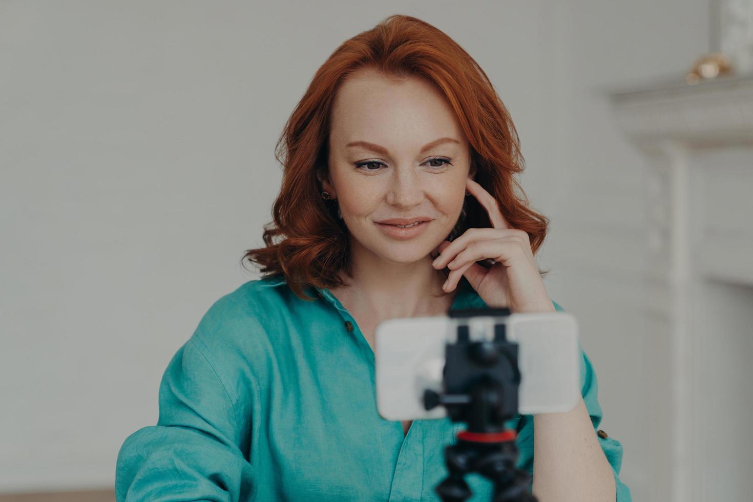 Photo of positive redhead woman blogger influencer shoots video for personal blog, uses modern mobile phone on tripod, does online chat, shoots blog at home, uses high speed internet connection