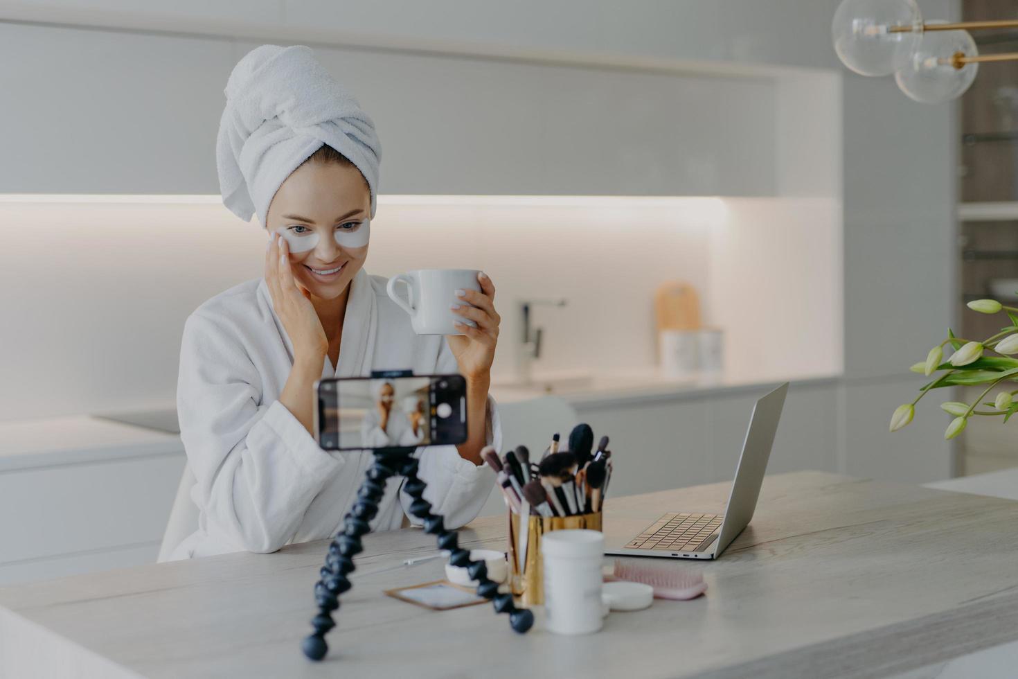 Woman beauty blogger shoots cosmetic vlog looks at camera of smartphone talks with followers applies collagen patches under eyes drinks tea wears white soft bathrobe records video broadcast. photo