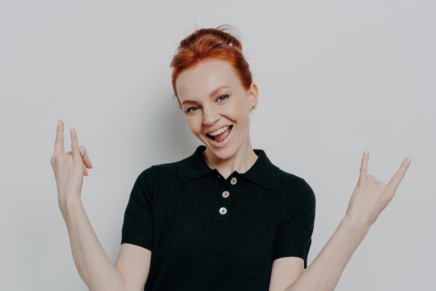 Confident young redhead woman doing rock and roll sign while standing isolated over grey background photo