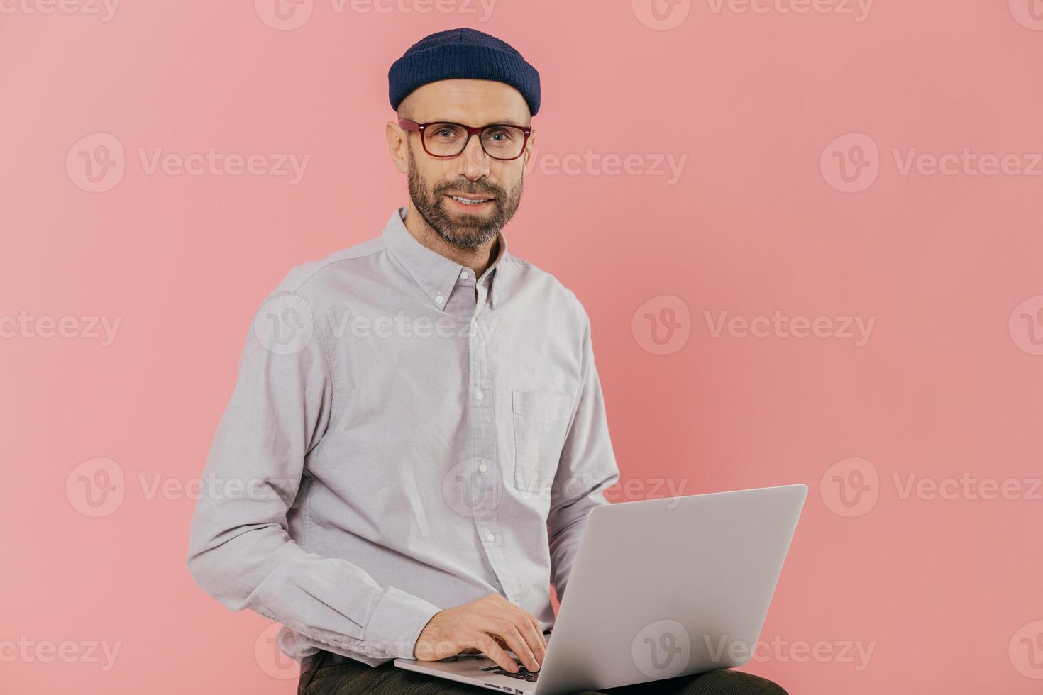 Male graphic designer thinks about creative ideas, reads news on internet website, uses moderm laptop computer, sits against pink background. Businessman reads publication online, makes research photo