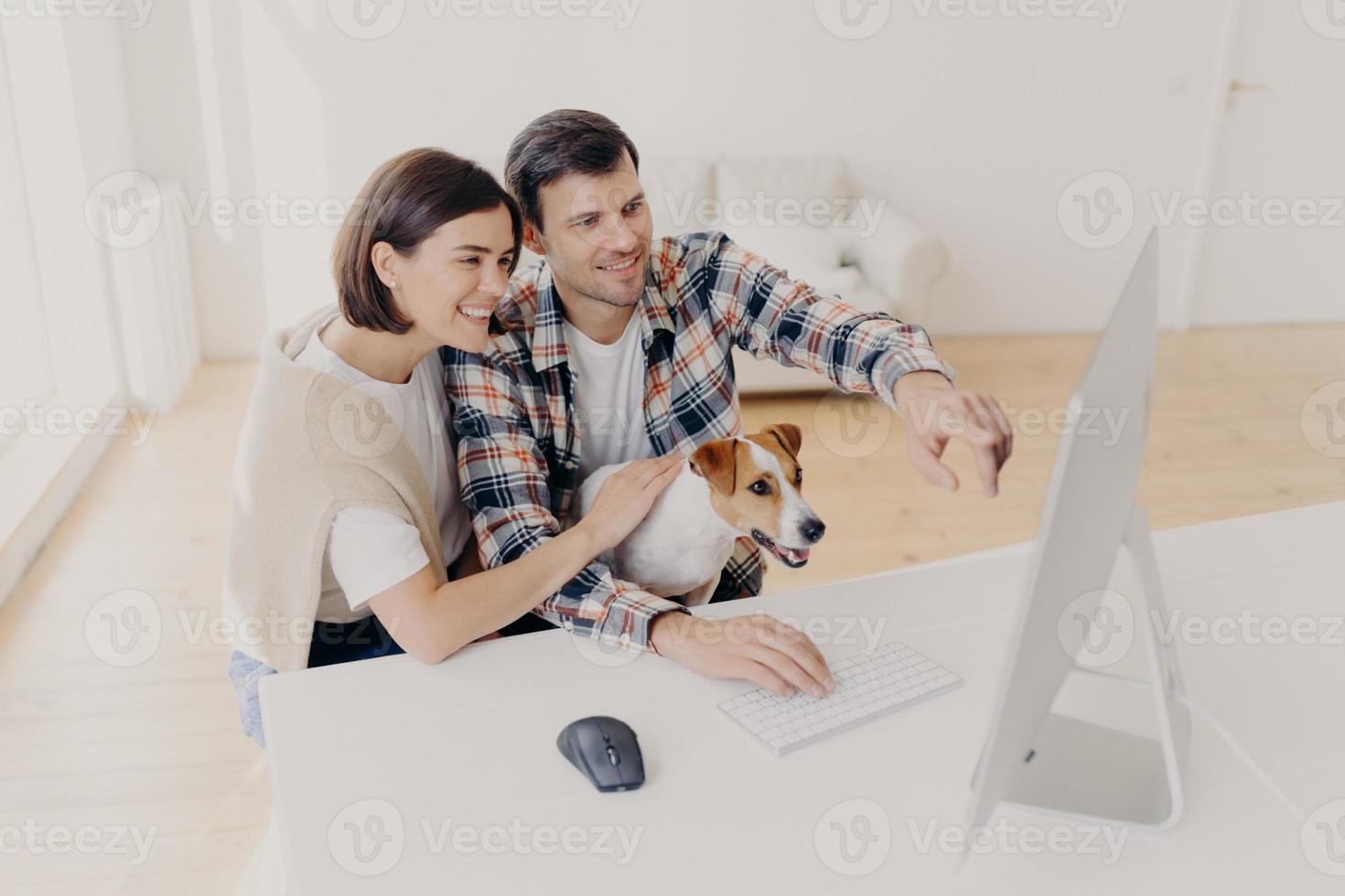 Overjoyed woman and man has happy looks, watch online movie at home together, play with dot, sit at white table, search information via wifi, dressed in casual clothes, Family couple browse internet photo
