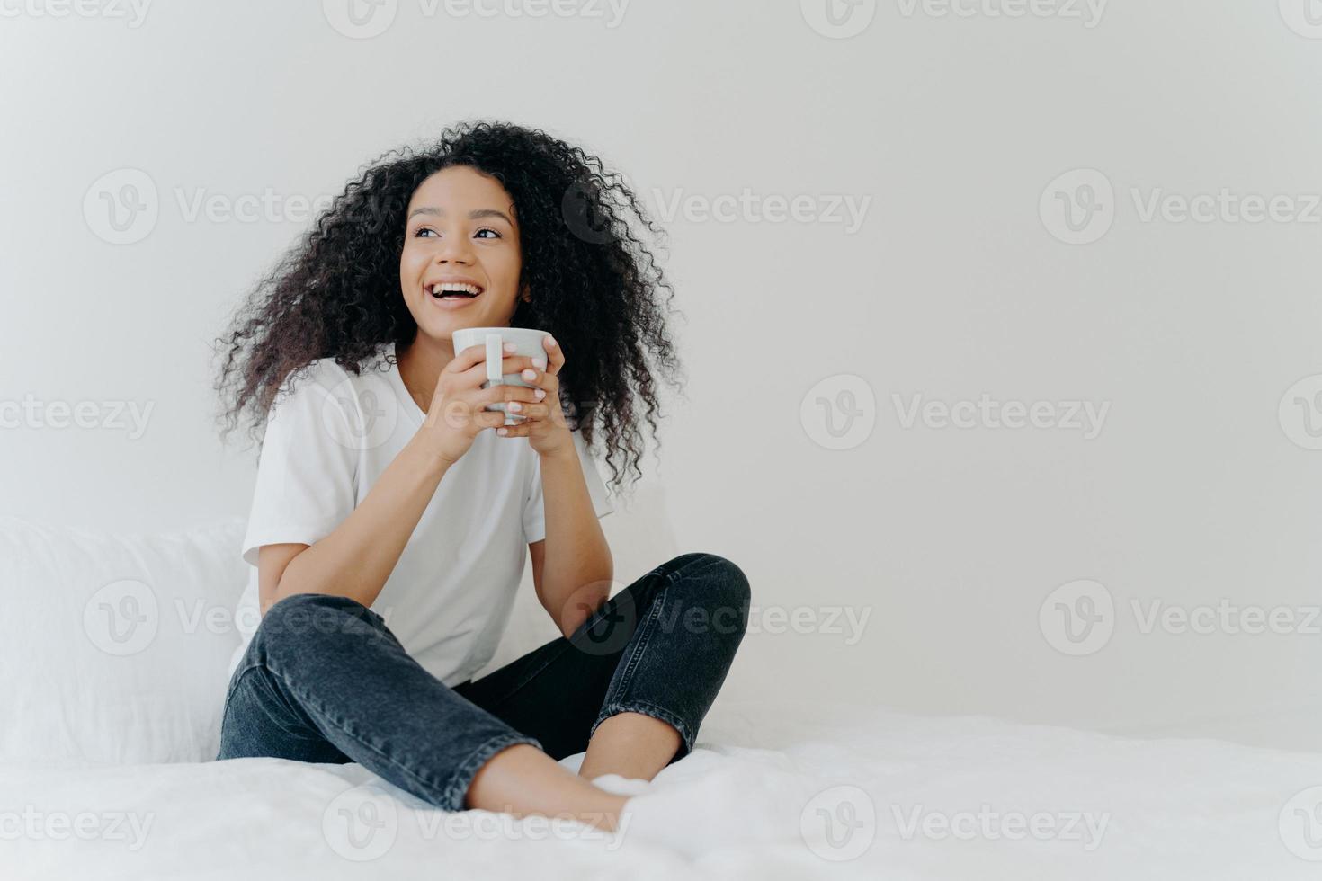 Lazy morning and bedtime concept. Positive Afro woman holds mug of hot tea or coffee, sits in bed, looks gladfully aside, enjoys good weather, wears comfortable clothes, white wall in background photo