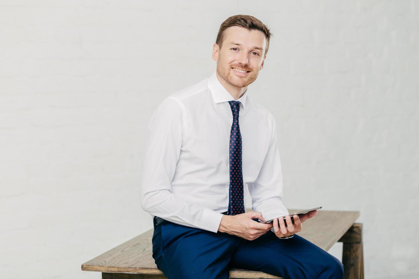 un joven y apuesto empleado vestido elegantemente, tiene un panel táctil moderno, piensa en el informe financiero y se reúne con socios comerciales, se sienta a la mesa, posa en el interior. concepto de personas y ocupación foto