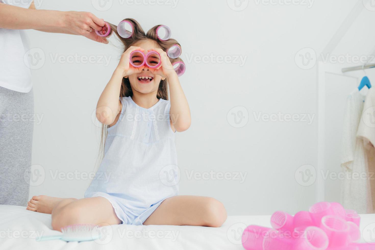 feliz linda niña se divierte, mantiene dos rulos cerca de los ojos, usa ropa de dormir, su madre enrolla rulos en su cabello largo, posa en una habitación blanca. concepto de niños, belleza, felicidad y alegría. foto