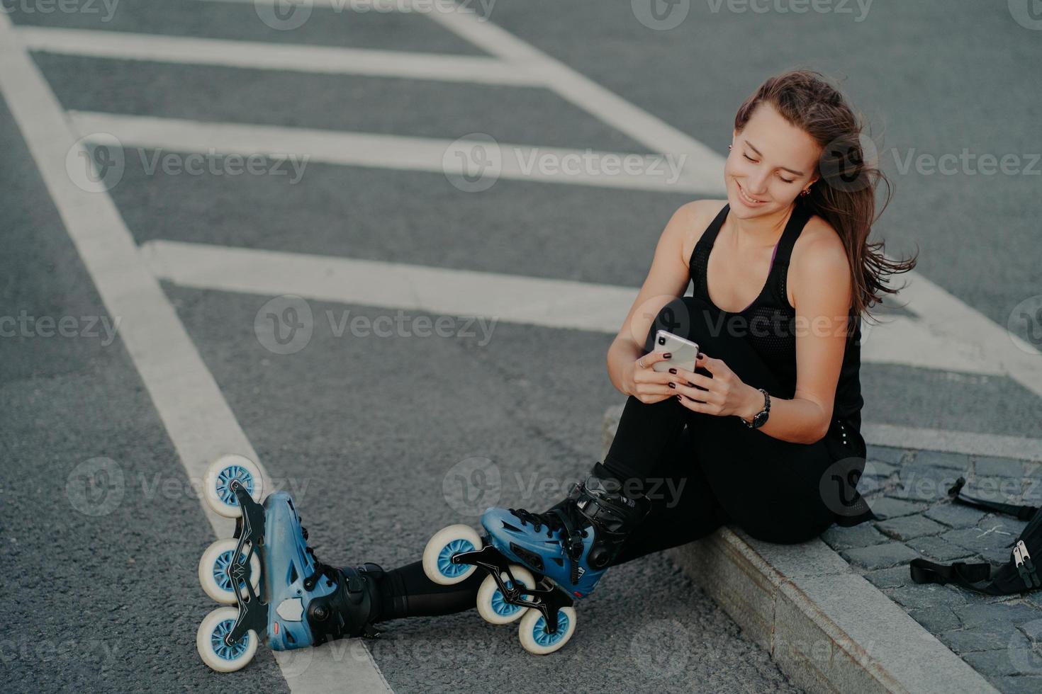 Top view of brunette active young woman uses smartphone for online communication poses on asphalt dressed in black active wear enjoys favorite hobby and free time. Extreme sport and rest concept photo