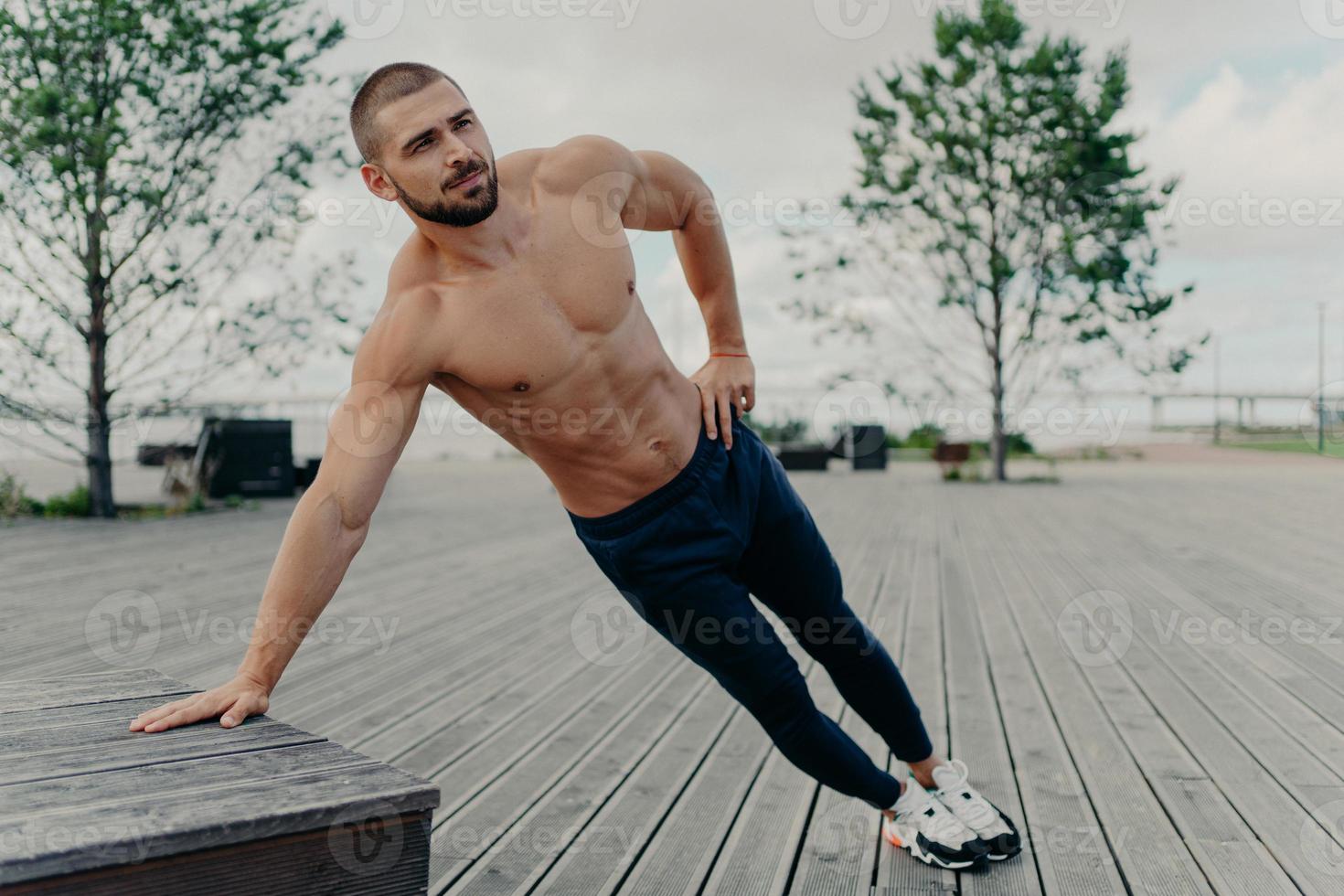 Young handsome shirtless ordinary bearded man stands in diagonal body position, makes side plank, practices endurance and stays in good physical shape. Sportsman in active wear trains outdoor photo