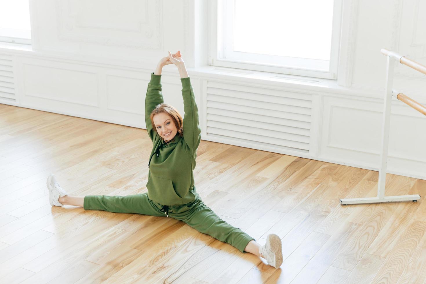 Una joven atleta pelirroja satisfecha practica yoga, hace ejercicios divididos, usa ropa deportiva y zapatos deportivos blancos, levanta las manos, demuestra buena flexibilidad, se sienta en el suelo cerca de la barra de ballet. foto