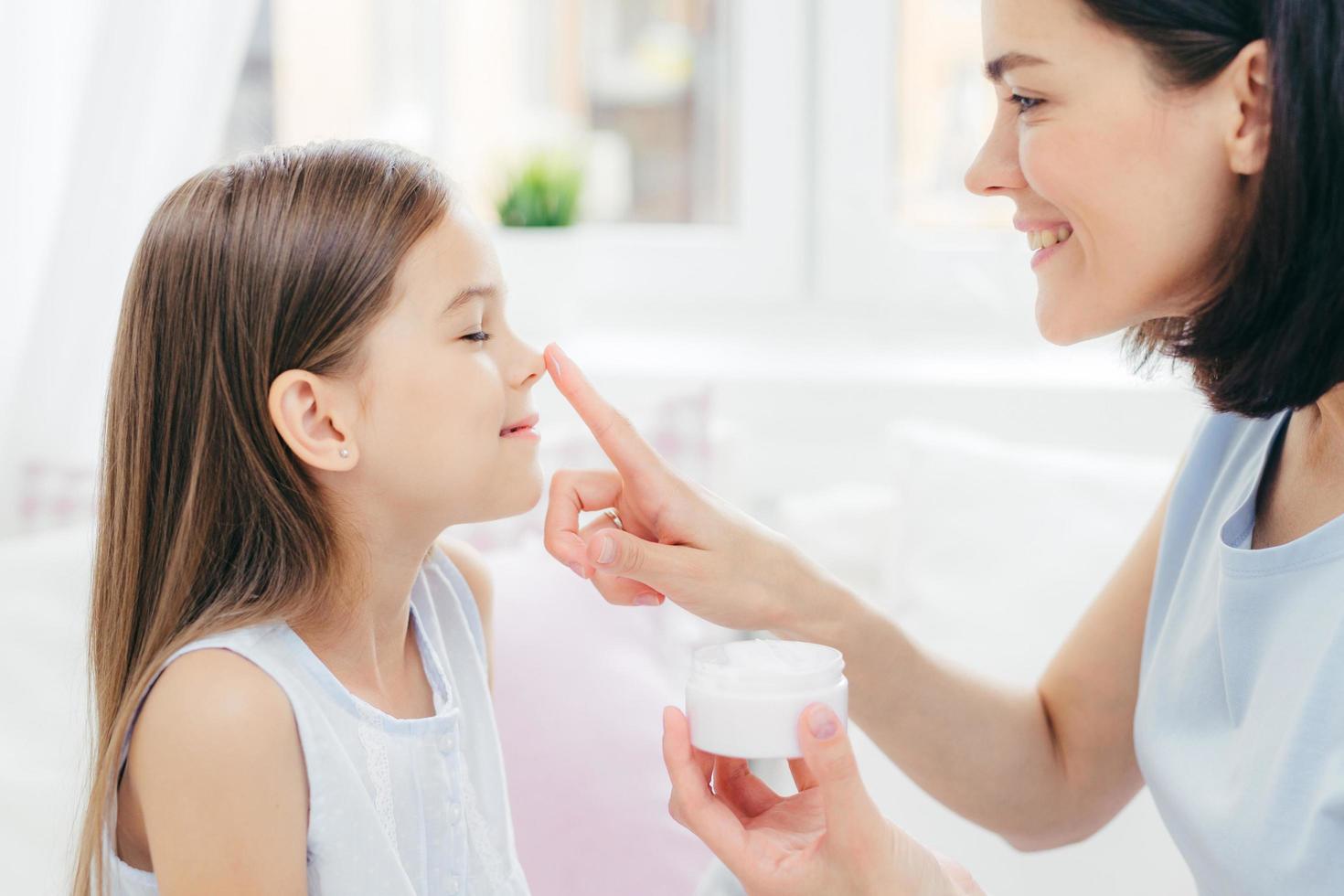 Cropped shot of affectionate young mum touches daughters nose, holds bottle of cream, enjoy togetherness, take care of skin. Beautiful happy woman with her child. People and cosmetics concept photo