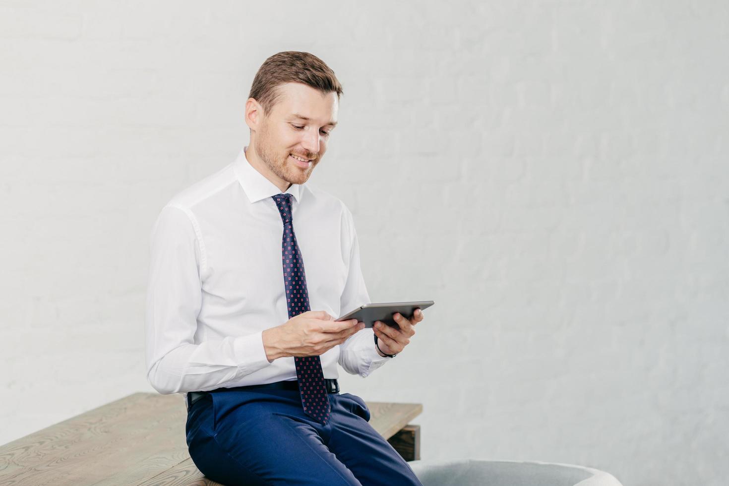 el financiero masculino positivo usa camisa blanca, corbata, se sienta en la mesa, instala una nueva aplicación en el panel táctil, conectado a Internet inalámbrico. exitoso hombre de negocios lee la notificación en un dispositivo moderno foto