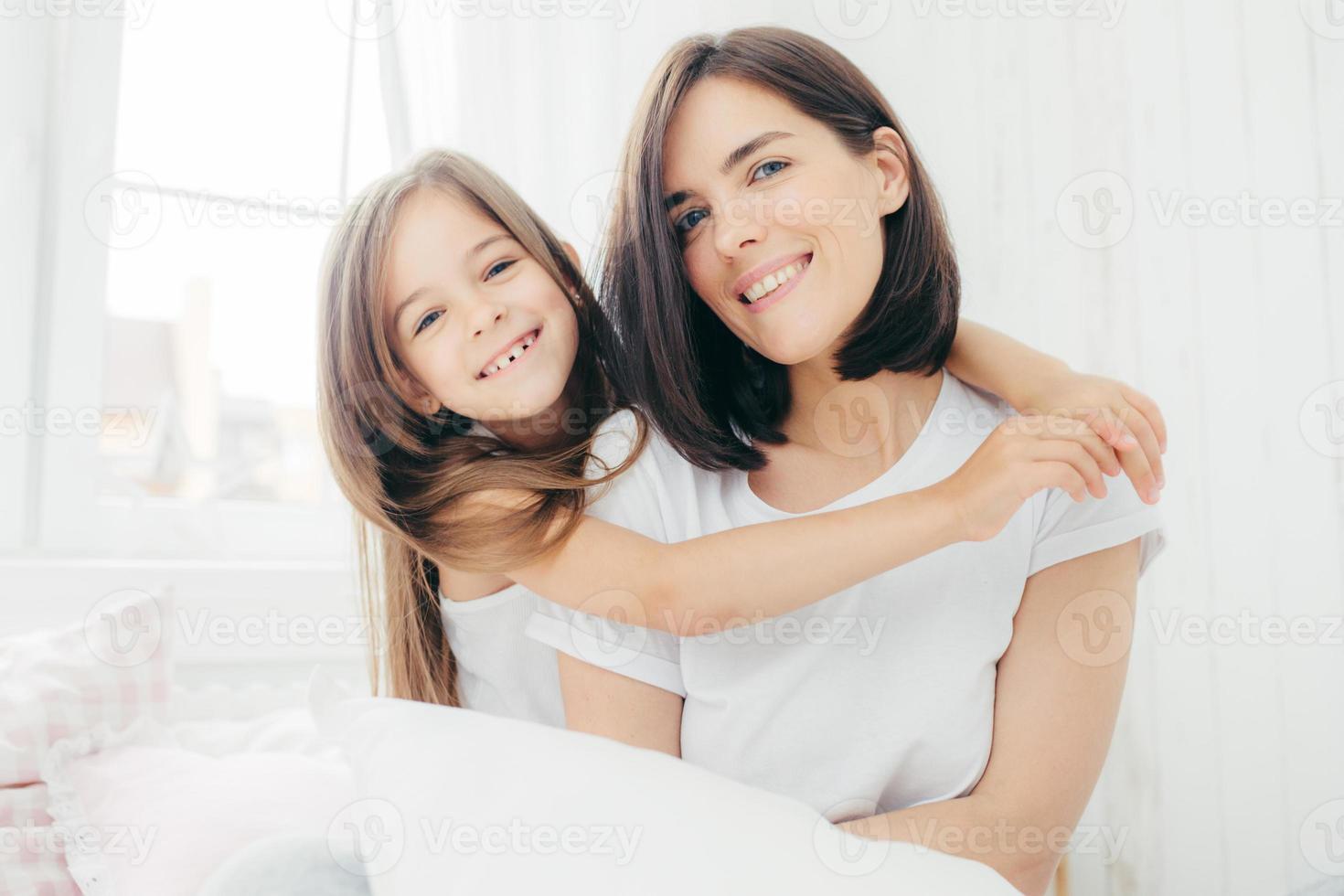 Portrait of happy Caucasian mother with charming smile and her small daughter embraces with love mum, being in good mood, pose on comfortable bed against cozy interior. People and family concept photo
