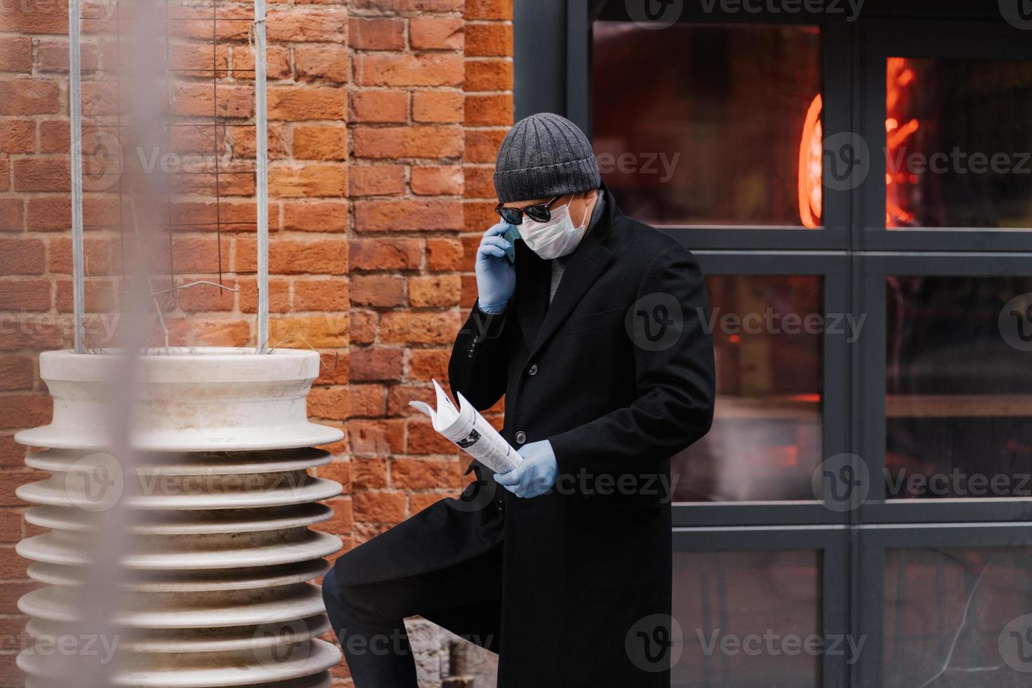 Male entrepreneur discusses working issuses via smartphone, wears protective medical mask and gloves, holds papers, poses brick wall. Epidemic virus. Novel coronavirus concept. Stay safe and healthy photo