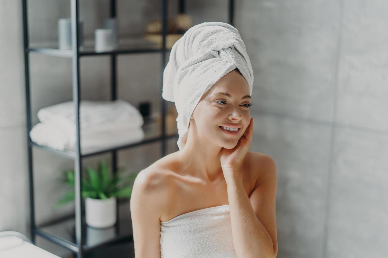 Positive European woman smiles pleasantly, touches healthy skin, wears wrapped towel on head, poses in bathroom. Spa lady with natural makeup poses refreshed after taking shower. Pampering, wellness photo
