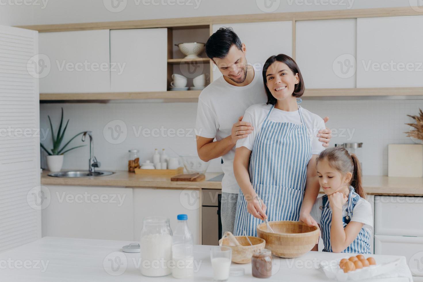 Happy family cook together at kitchen. Father, mother and dauther busy preparing delicious meal at home. Husband embraces wife who whisks and prepares dough, bake cookies. Food, togetherness photo