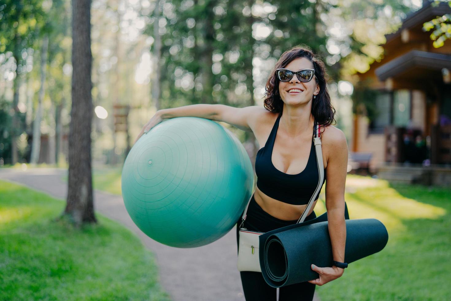 mujer alegre sonríe ampliamente, sostiene una gran pelota de fitness, karemat enrollado, usa gafas de sol, disfruta el día de verano, practica deporte regularmente, es instructora de pilates, posa al aire libre en la hermosa naturaleza foto