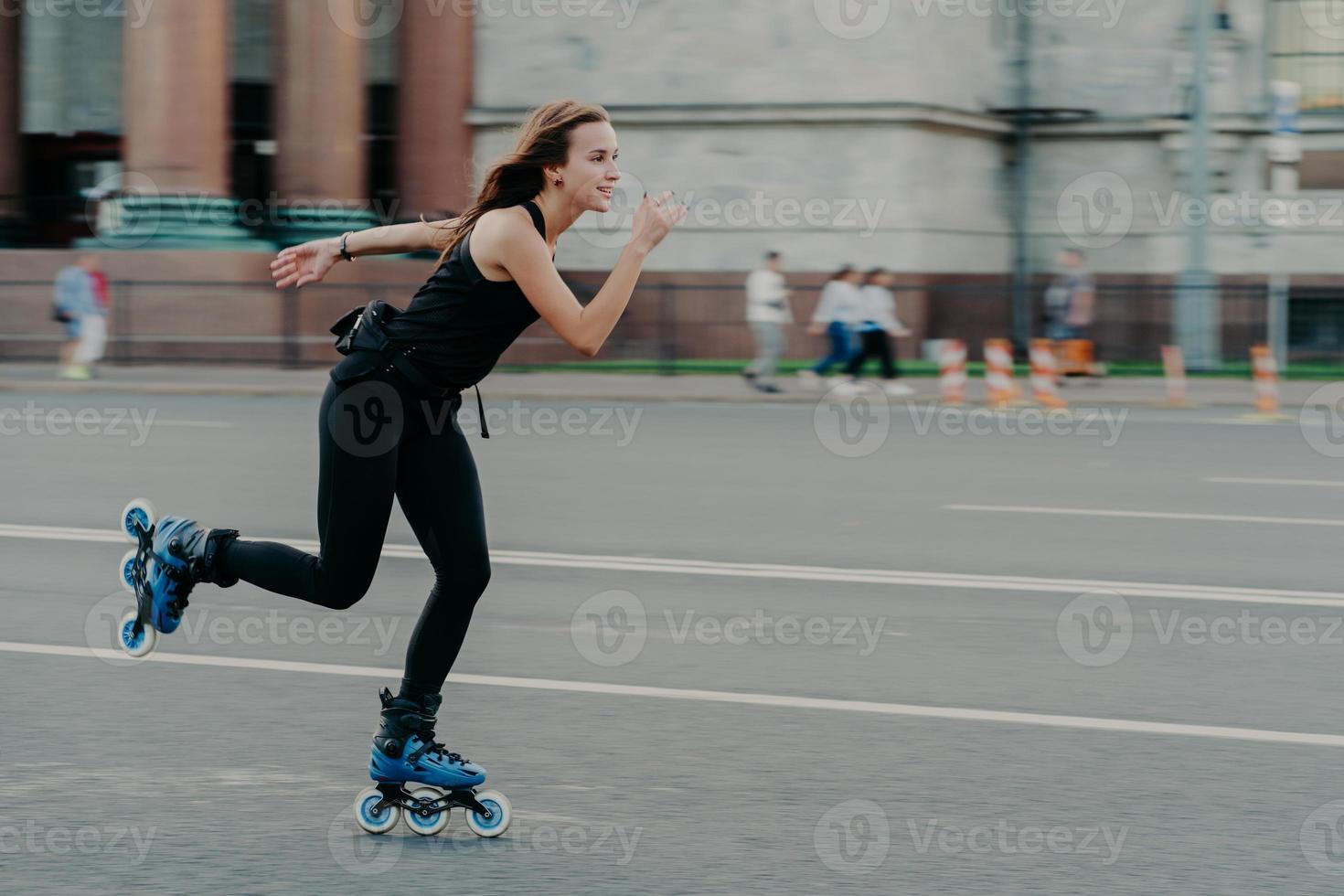 Holidays and active lifestyle concept. Slim healthy European woman rollerskates on high speed photogaphed in motion dressed in black clothing poses against blurred street background on road. photo