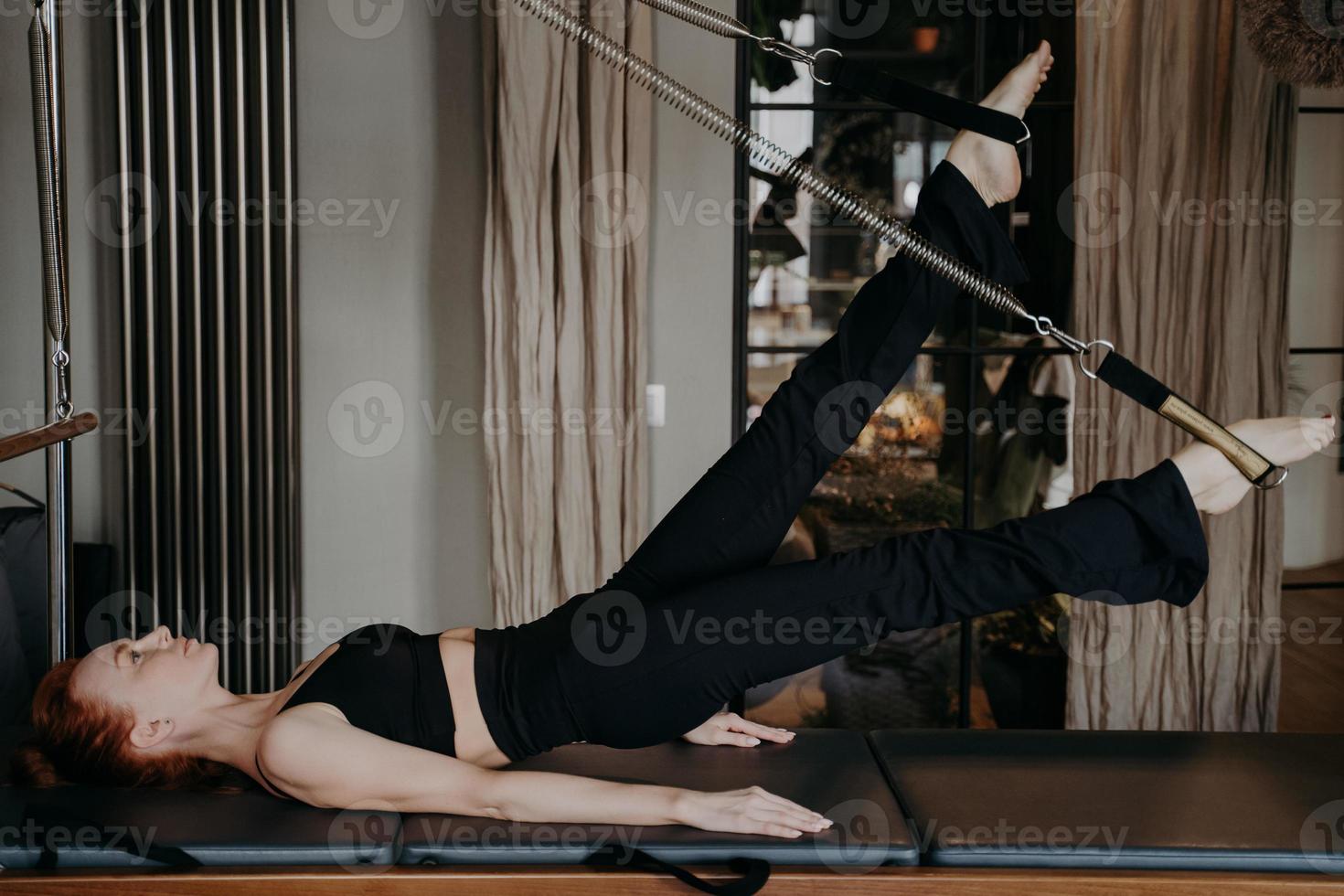 mujer pelirroja flexible haciendo ejercicios de estiramiento de piernas en cadillac reformer foto