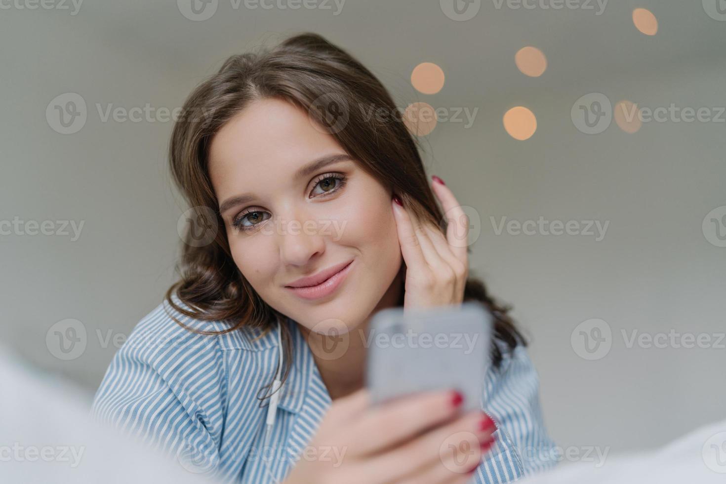 hermosa mujer caucásica escucha audio con auriculares, conectada a un moderno dispositivo de teléfono inteligente, ve videos en las redes sociales, yace en la cama en el interior. concepto de personas, tecnología y entretenimiento foto