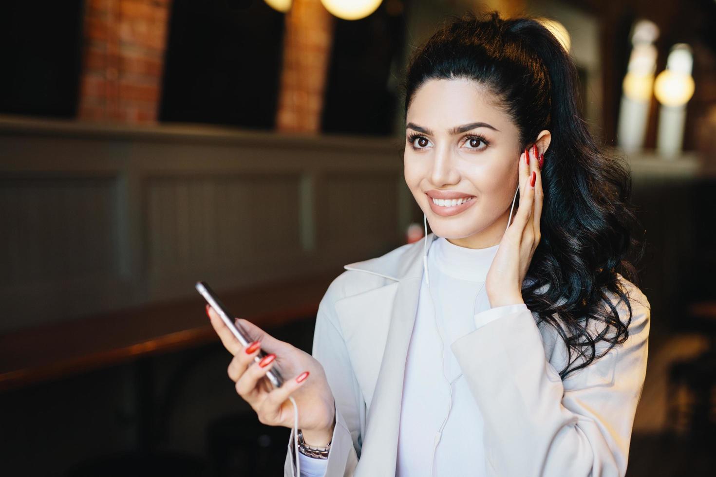 Beautiful brunette woman with expressive eyes and long eyelashes, having make-up and red manicure wearing white jacket listening to her favourite audio book having happy expression sitting indoors photo