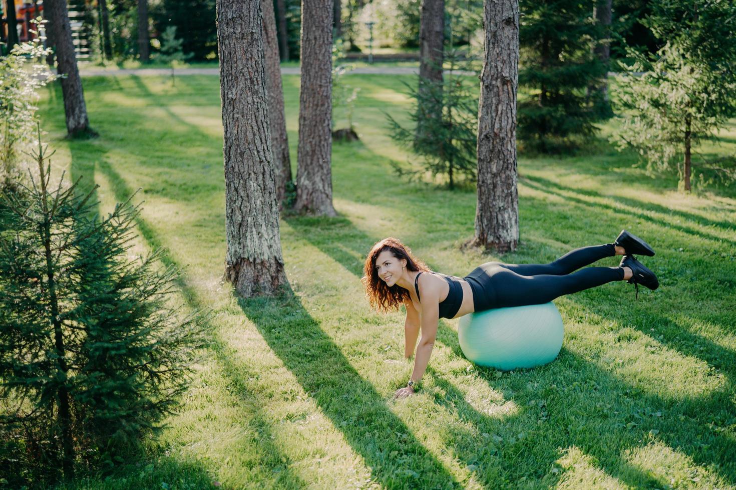 Above view of determined young brunette European woman exercises pilates in green park, balances on fitness ball, dressed in activewear, looks happily somewhere poses outside. Selective focus photo