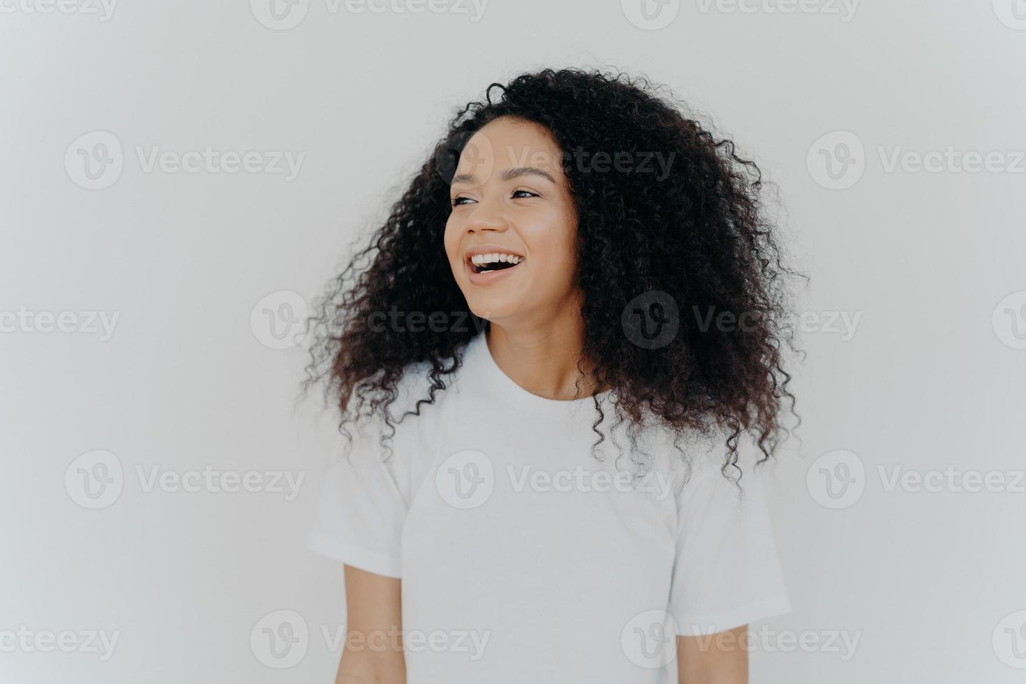 Portrait of beautiful curly adult woman turns gaze aside, has glad expression, smiles and laughs at something funny, dressed in white t shirt, models indoor, feels free, energized and relieved, photo