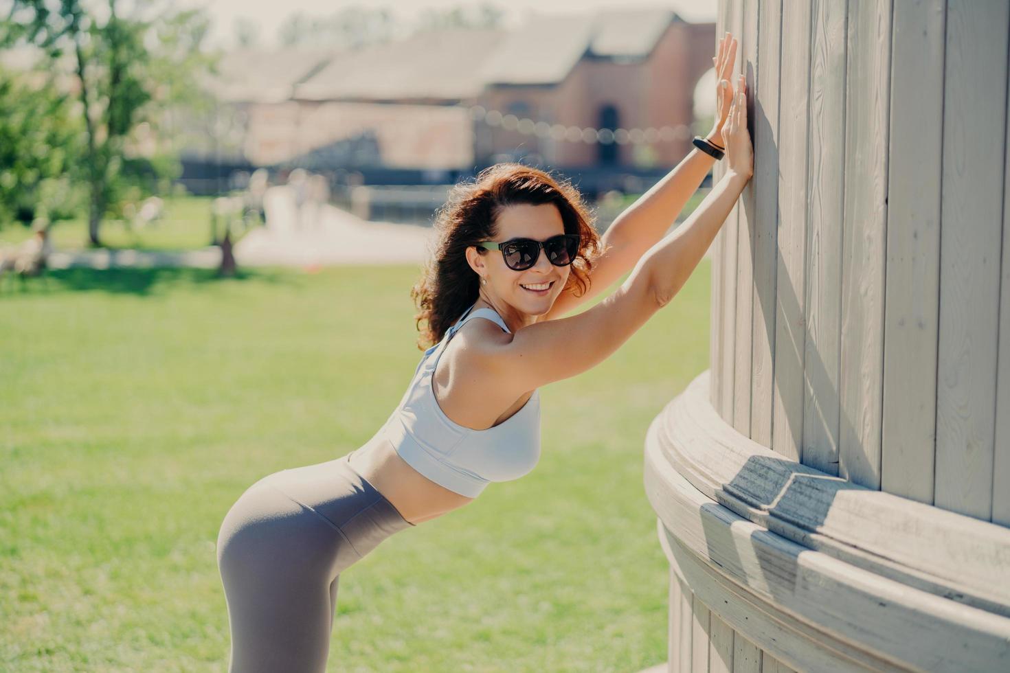 Slim woman with perfect body shapes leans with hands at something smiles positively dressed in top and leggings poses outdoor has fitness training during summer time. People and exercising concept photo