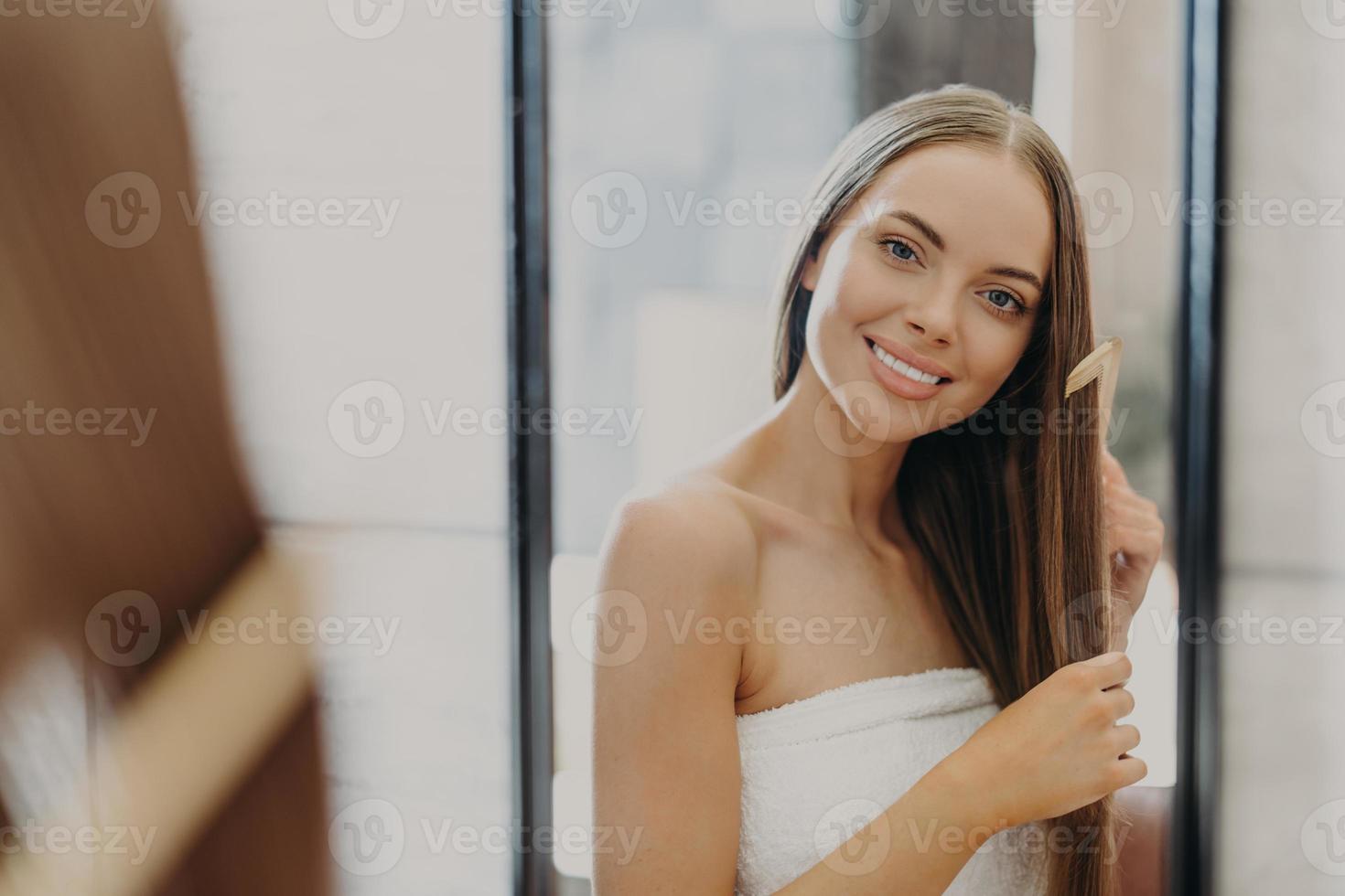 retrato de hermosa mujer hermosa con hombros desnudos peina su cabello lacio, sonríe ampliamente y mira su reflejo en el espejo, posa en el baño, envuelto en toalla. concepto de belleza de las mujeres foto